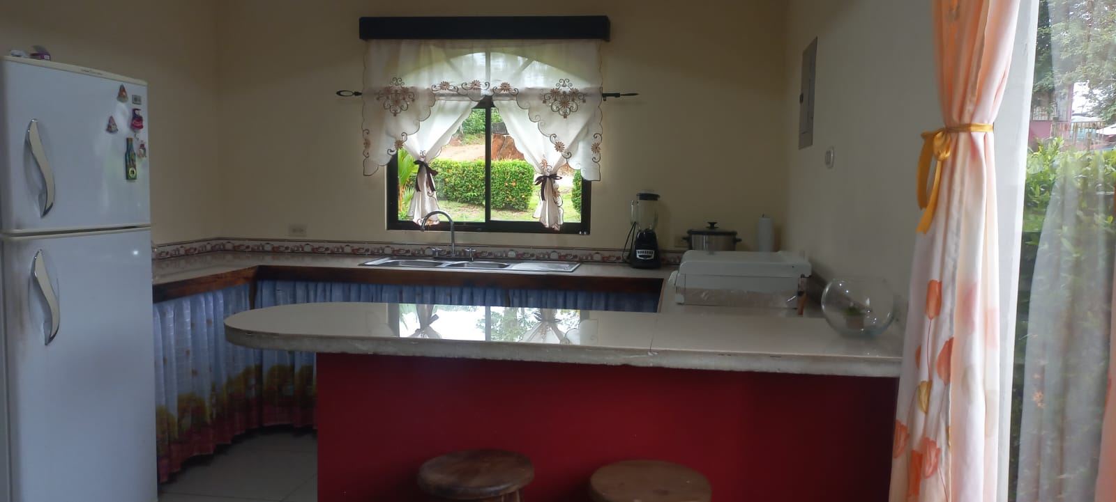 Kitchen with a white fridge on the left, sink under a window, and a red island with two wooden stools in the foreground.