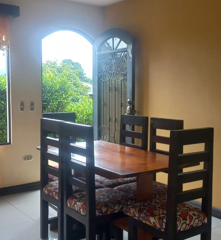 Wood dining table with four black chairs and colorful, patterned cushions in a sunlit dining area near an arched doorway to greenery outside.