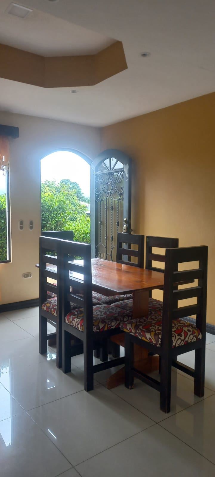 Wood dining table with four black chairs and colorful, patterned cushions in a sunlit dining area near an arched doorway to greenery outside.