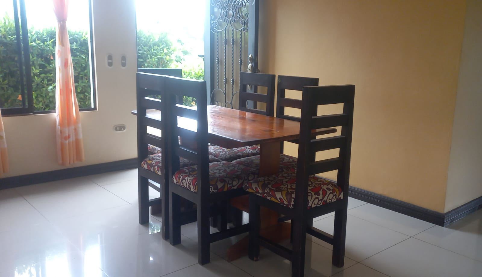 Wooden dining table with four black chairs and patterned cushioned seats in a bright room, peach curtains at a window, and greenery outside the open door area.