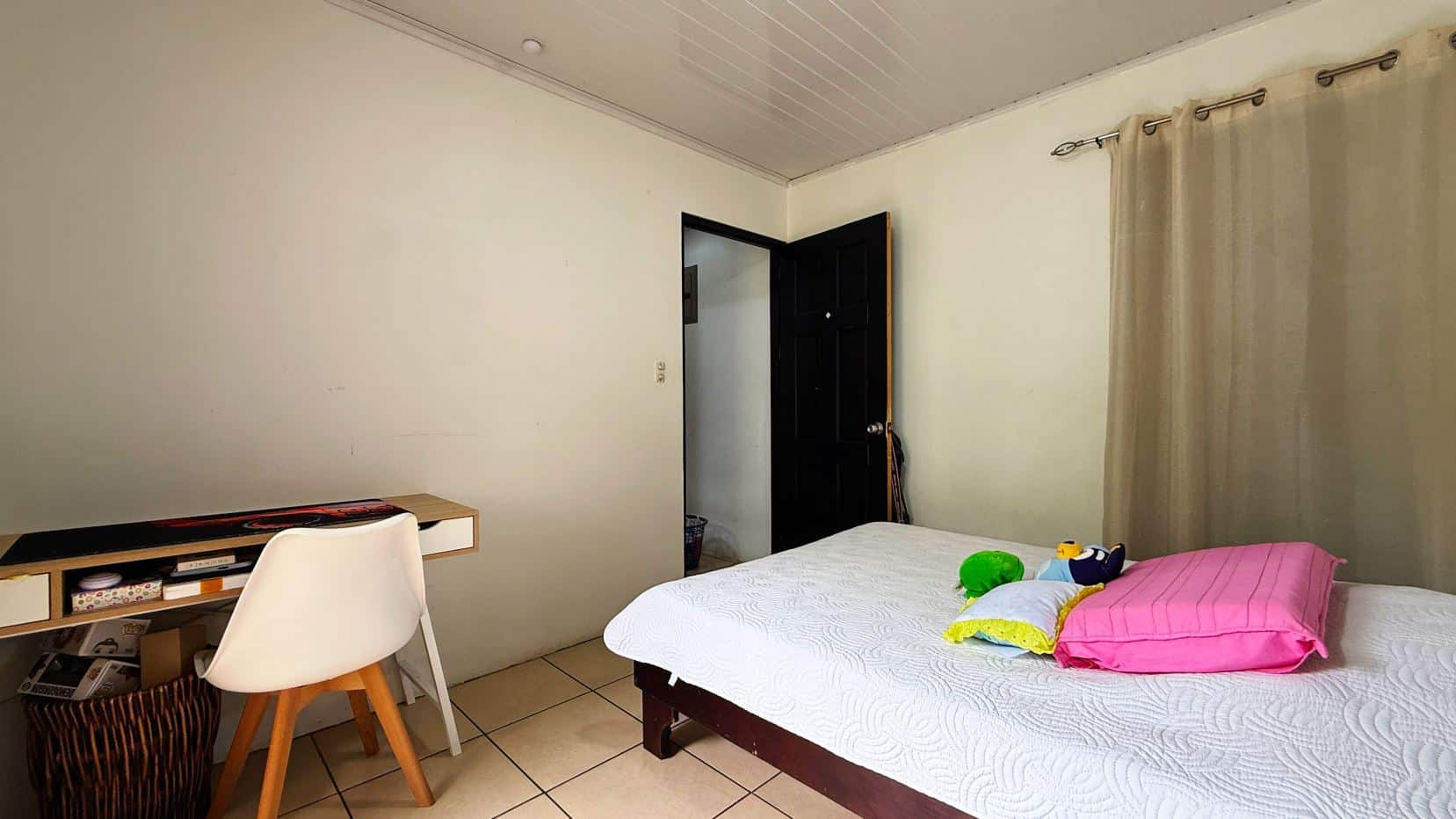 Bedroom with a bed on the right, pink pillows and stuffed toys, a desk and chair on the left, and an open door to another room.