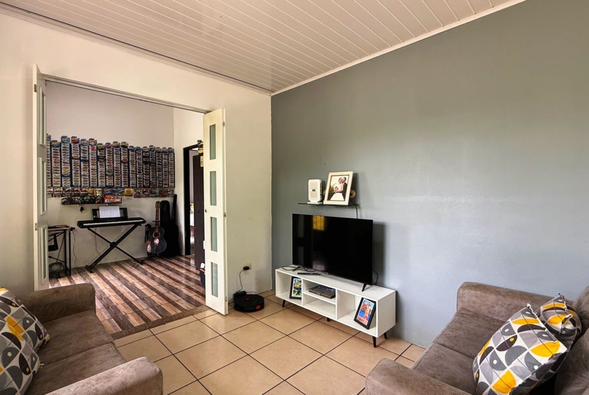 Living room scene with a gray accent wall, a TV on a white stand, and two patterned sofas facing inward; doorway reveals a room with shelves of model car collections and a keyboard.
