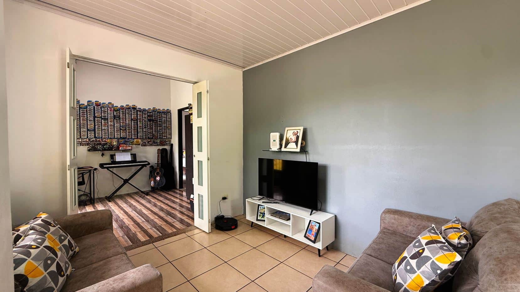 Living room scene with a gray accent wall, a TV on a white stand, and two patterned sofas facing inward; doorway reveals a room with shelves of model car collections and a keyboard.