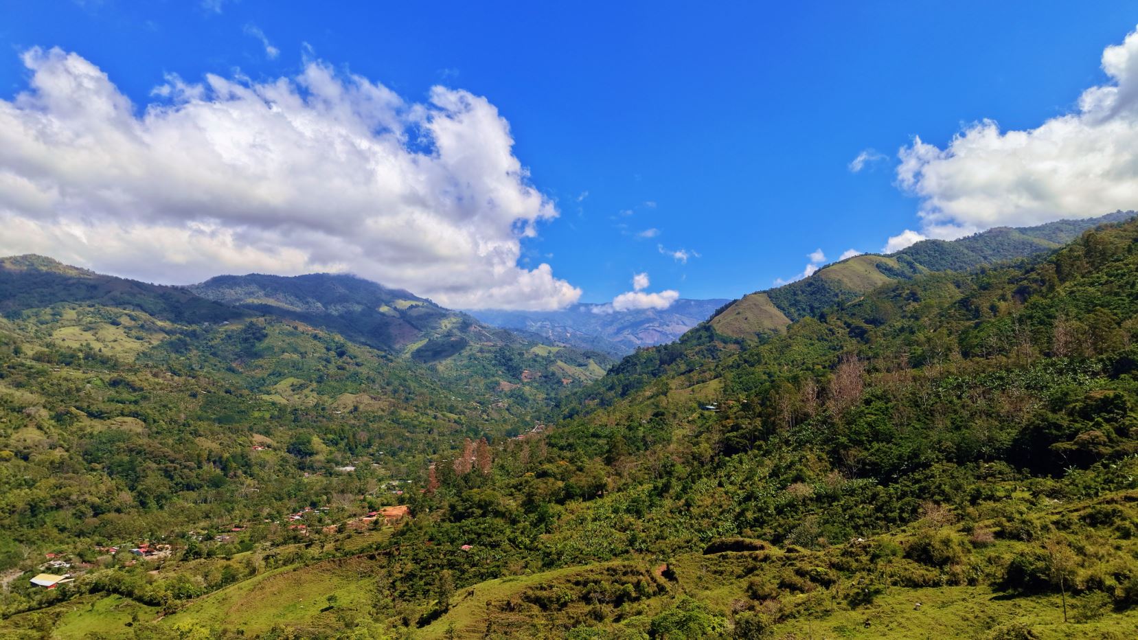 Wide green valley and rolling hills under a bright blue sky with fluffy white clouds; scattered houses dot the valley floor