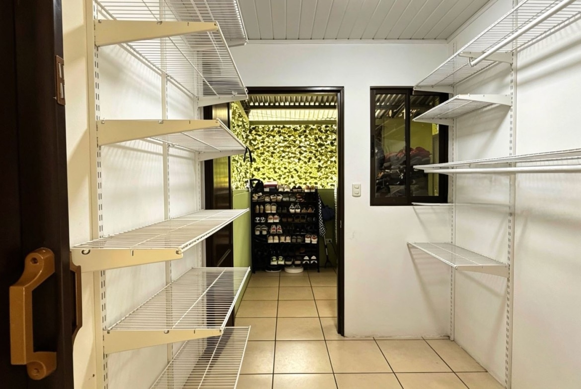 Narrow walk-in storage room with white metal shelving on both sides, leading to a back area with a shoe rack and plants on a green wall.