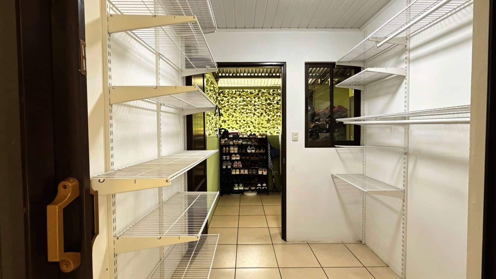 Narrow walk-in storage room with white metal shelving on both sides, leading to a back area with a shoe rack and plants on a green wall.