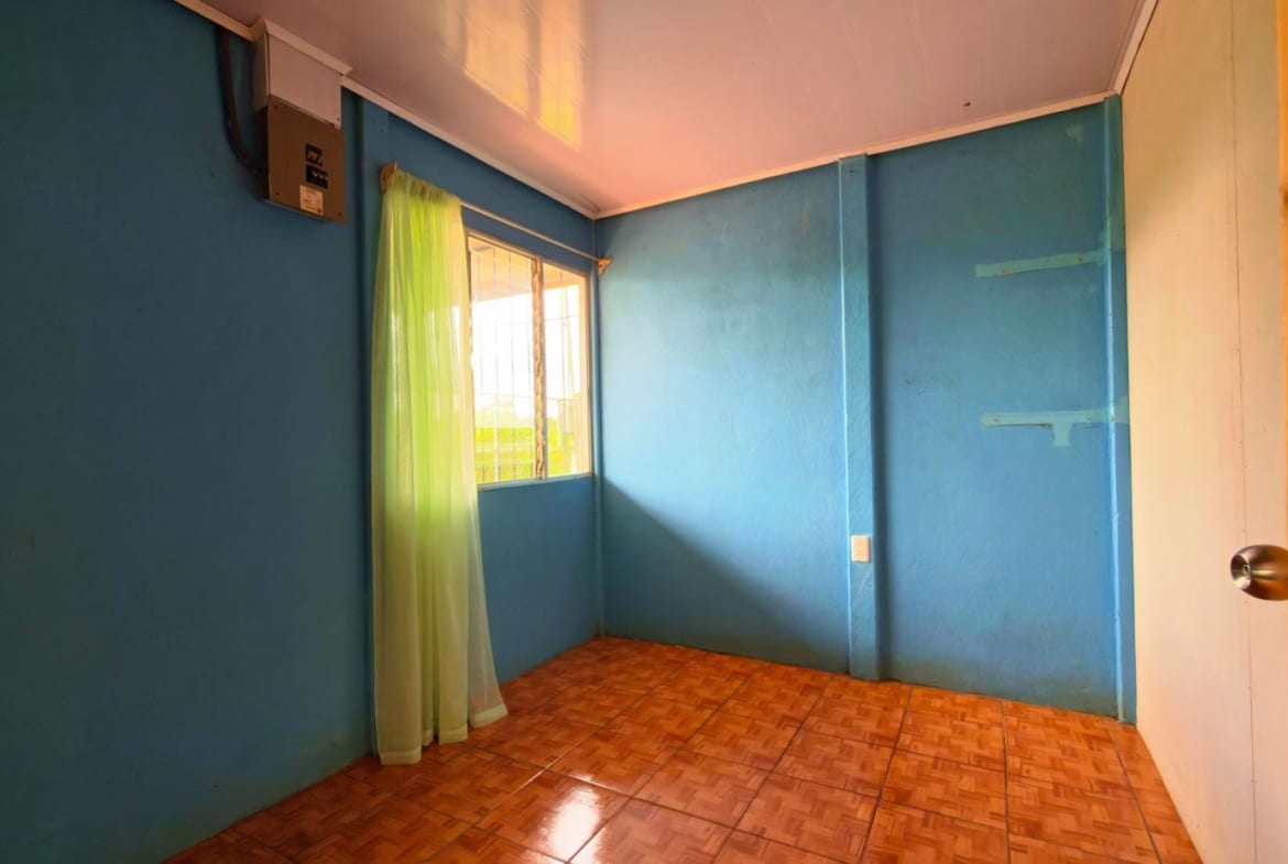 Small blue-painted room with brown tiled floor and a window with a sheer green curtain letting in sunlight.