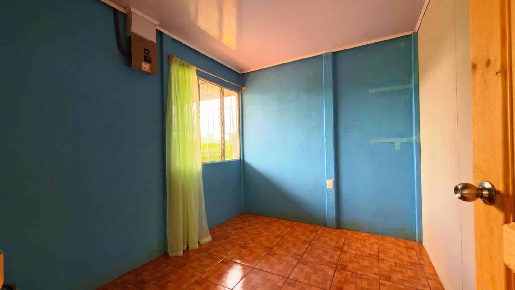 Small blue-painted room with brown tiled floor and a window with a sheer green curtain letting in sunlight.