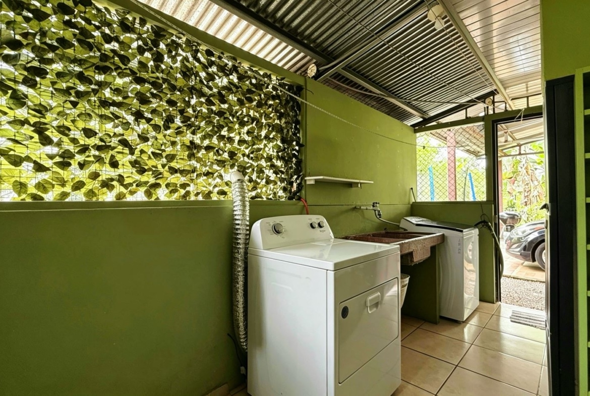 Green laundry room featuring a white front-loading washer and dryer, a utility sink, and open doorway to an outdoor area with a car visible outside. The wall has leafy lattice window and green shelving.