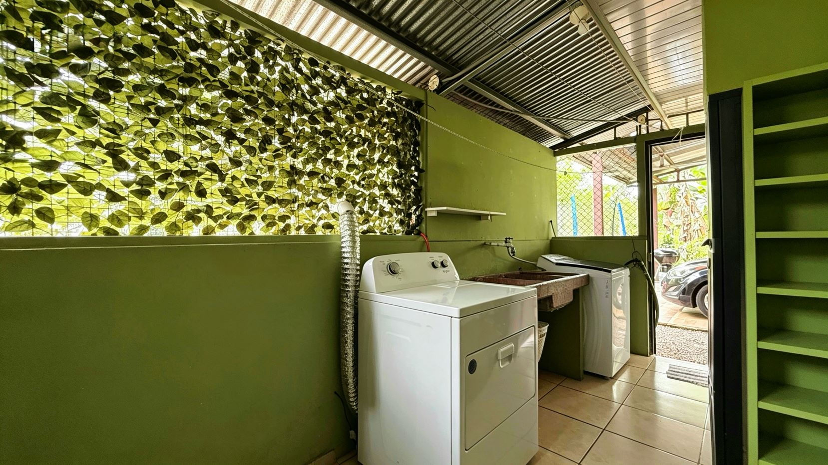 Green laundry room featuring a white front-loading washer and dryer, a utility sink, and open doorway to an outdoor area with a car visible outside. The wall has leafy lattice window and green shelving.