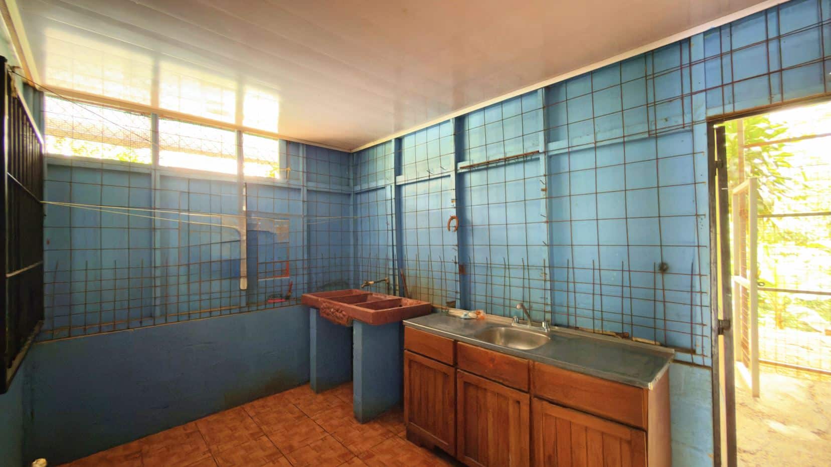 Blue tiled utility kitchen with a double sink, wooden cabinets, and sunlight streaming through an open door exterior view