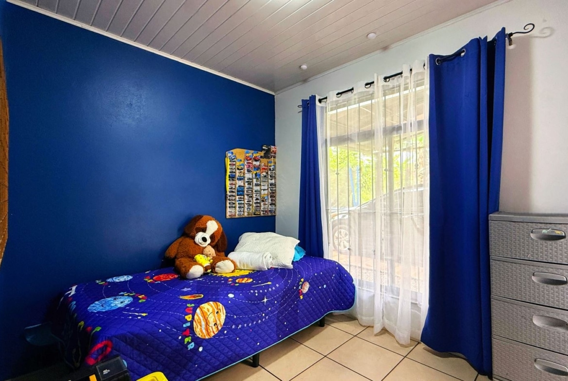 Child's bedroom with a blue accent wall, a single bed in space-themed quilt and stuffed animal, and a window with white and blue curtains beside a grey dresser.