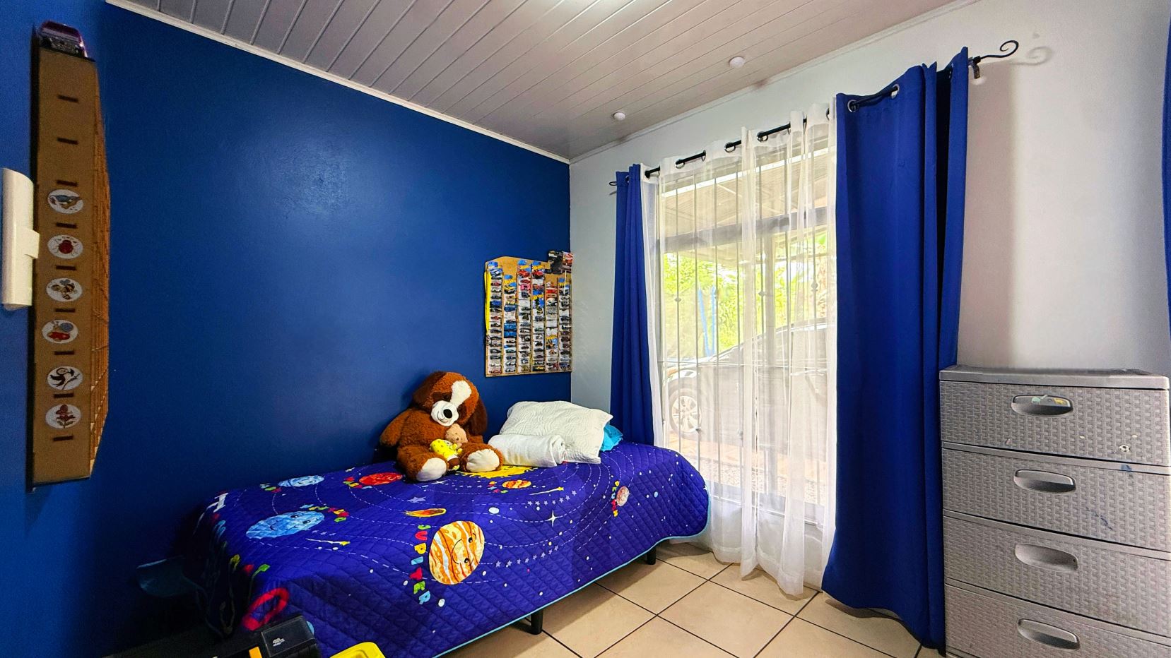 Child's bedroom with a blue accent wall, a single bed in space-themed quilt and stuffed animal, and a window with white and blue curtains beside a grey dresser.