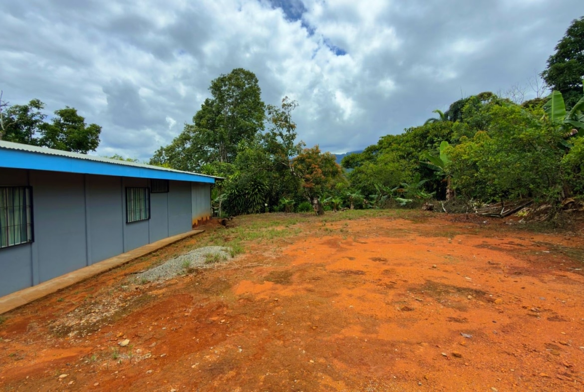 Single-story gray building with a blue roof on a red dirt yard, surrounded by green tropical trees under a cloudy sky.