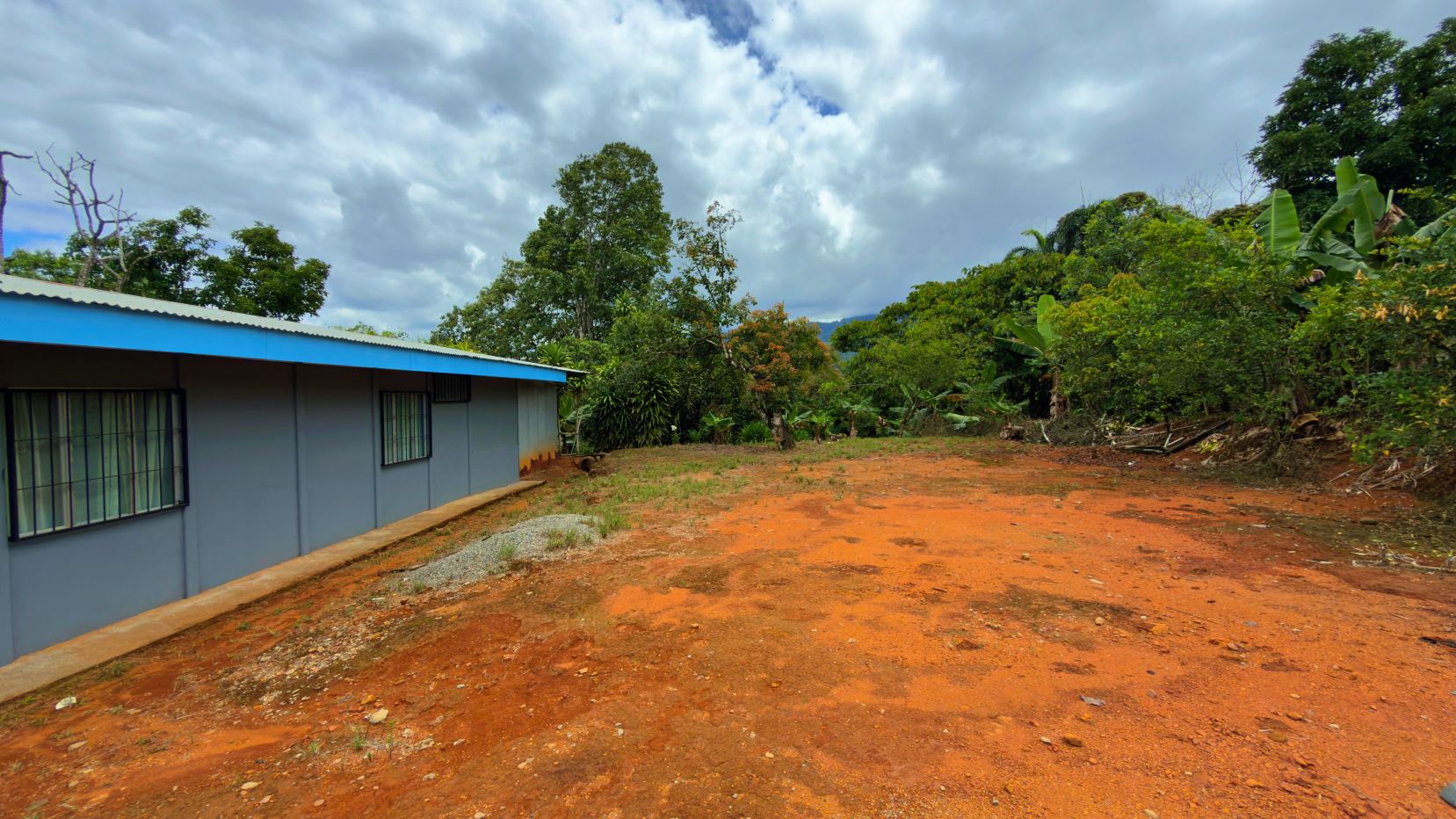 Single-story gray building with a blue roof on a red dirt yard, surrounded by green tropical trees under a cloudy sky.