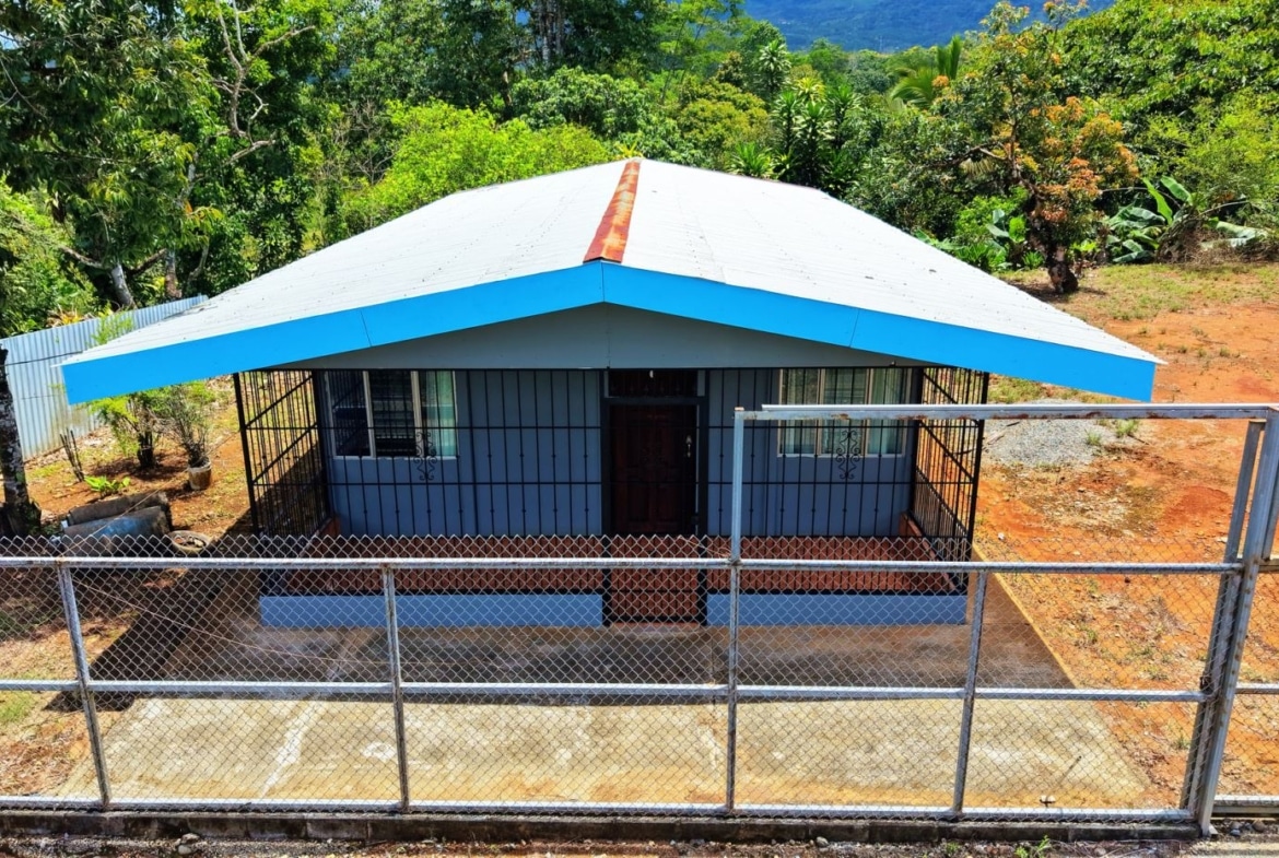 Front view of a small blue house with a white roof, fenced yard, and lush tropical trees in the background.