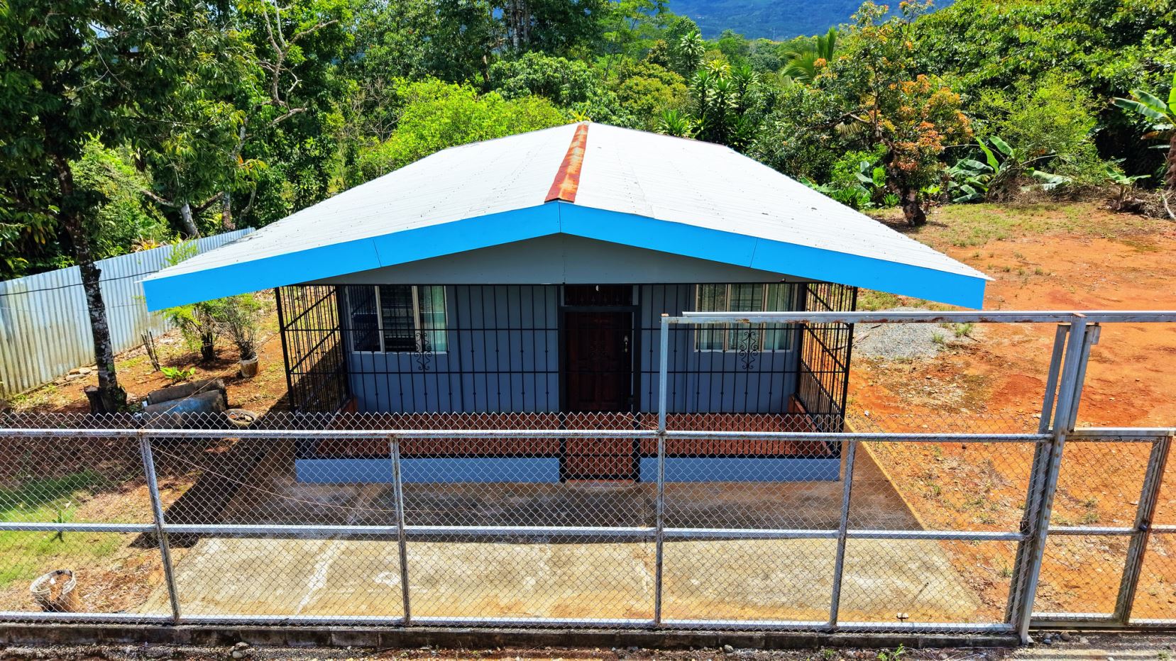 Front view of a small blue house with a white roof, fenced yard, and lush tropical trees in the background.