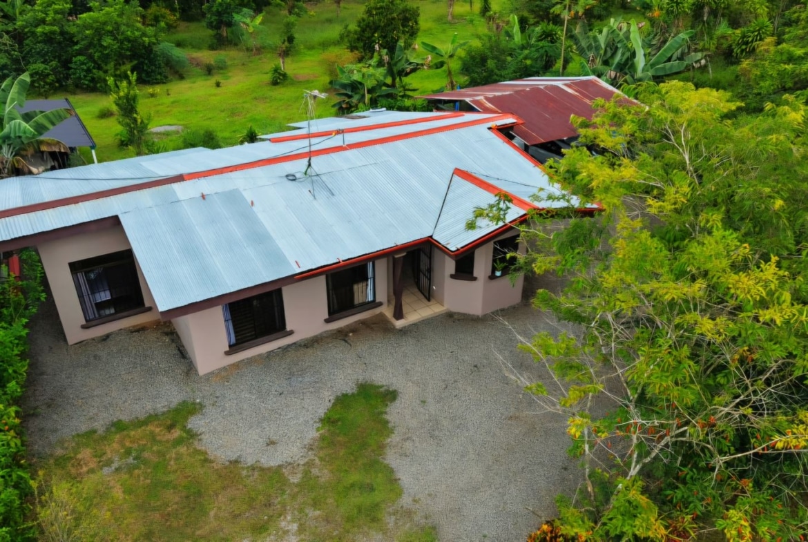 Aerial view of a single-story beige house with a silver corrugated metal roof edged in red, set in a lush tropical yard with gravel around it.