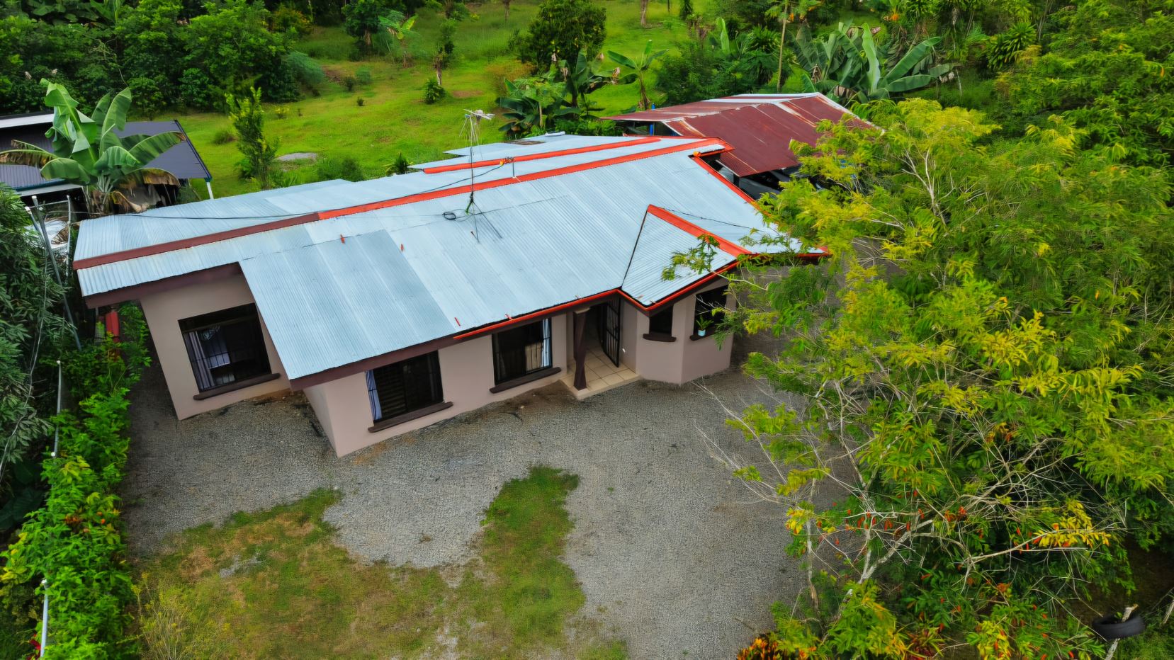 Aerial view of a single-story beige house with a silver corrugated metal roof edged in red, set in a lush tropical yard with gravel around it.