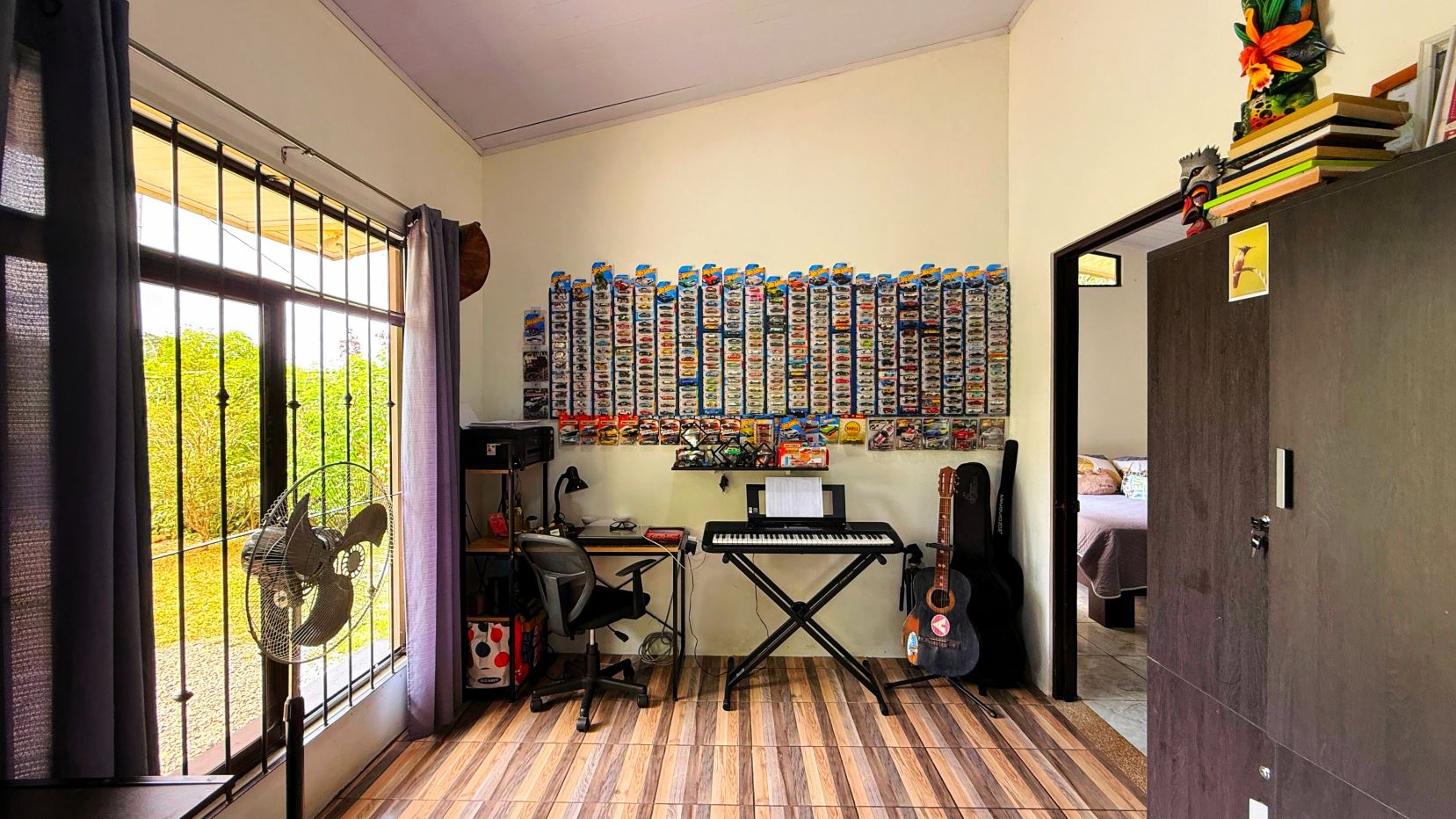 Room used as a small home studio: keyboard on a stand, guitar leaning against the wall, and a wall covered in rows of toy car package cards.
