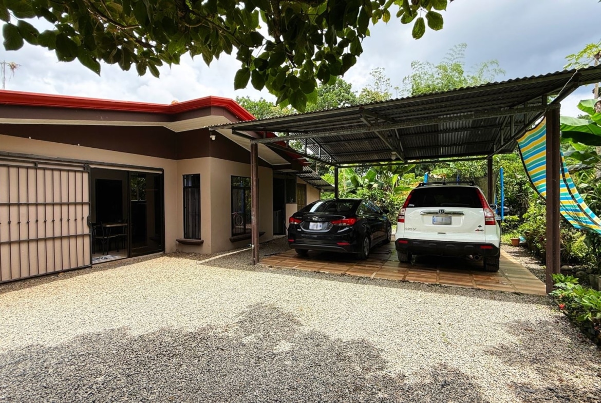 Single-story beige house with a metal carport holding a black sedan and a white SUV, gravel driveway and tropical plants nearby.