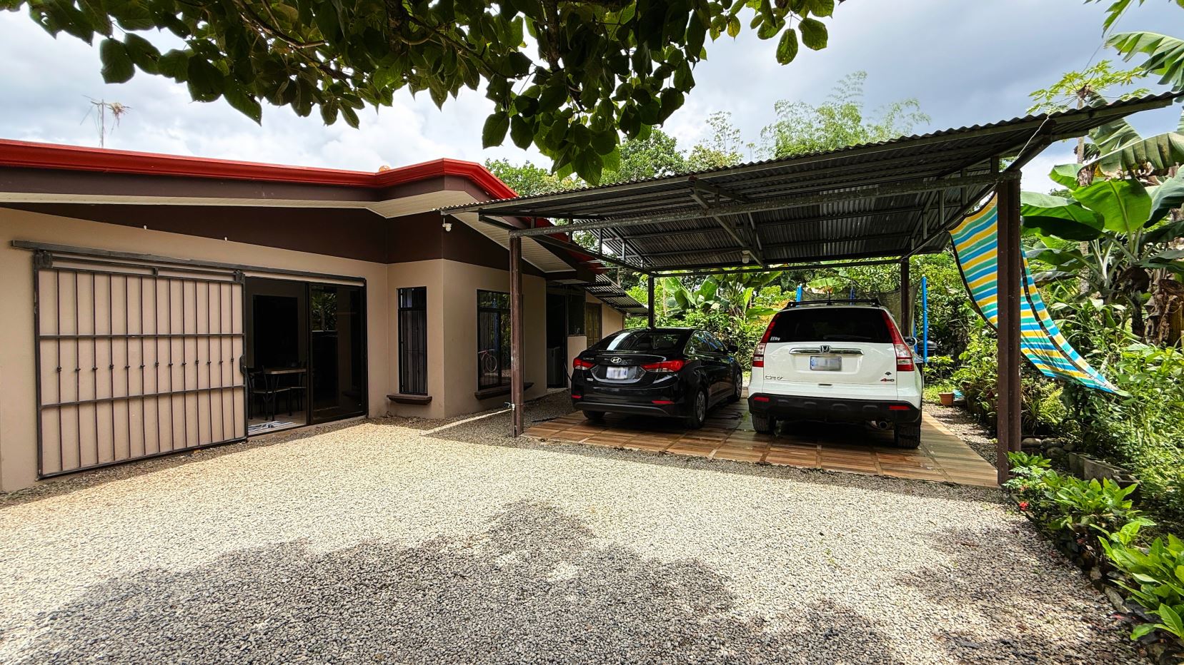 Single-story beige house with a metal carport holding a black sedan and a white SUV, gravel driveway and tropical plants nearby.