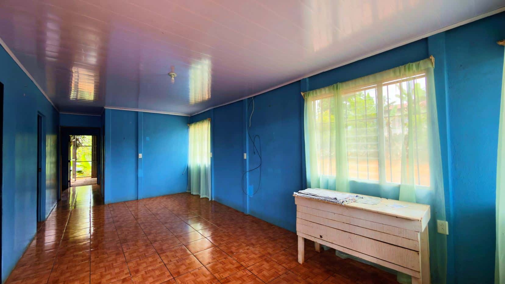 Blue-walled room with brown tiled floor, a large window with light green sheer curtains, and a white wooden dresser by the window outside view shows trees.