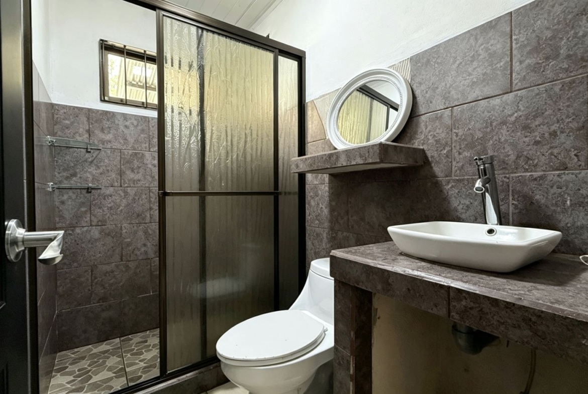 Bathroom with brown tile walls, frosted glass shower enclosure, white vessel sink on a brown countertop, round mirror, and a white toilet nearby.