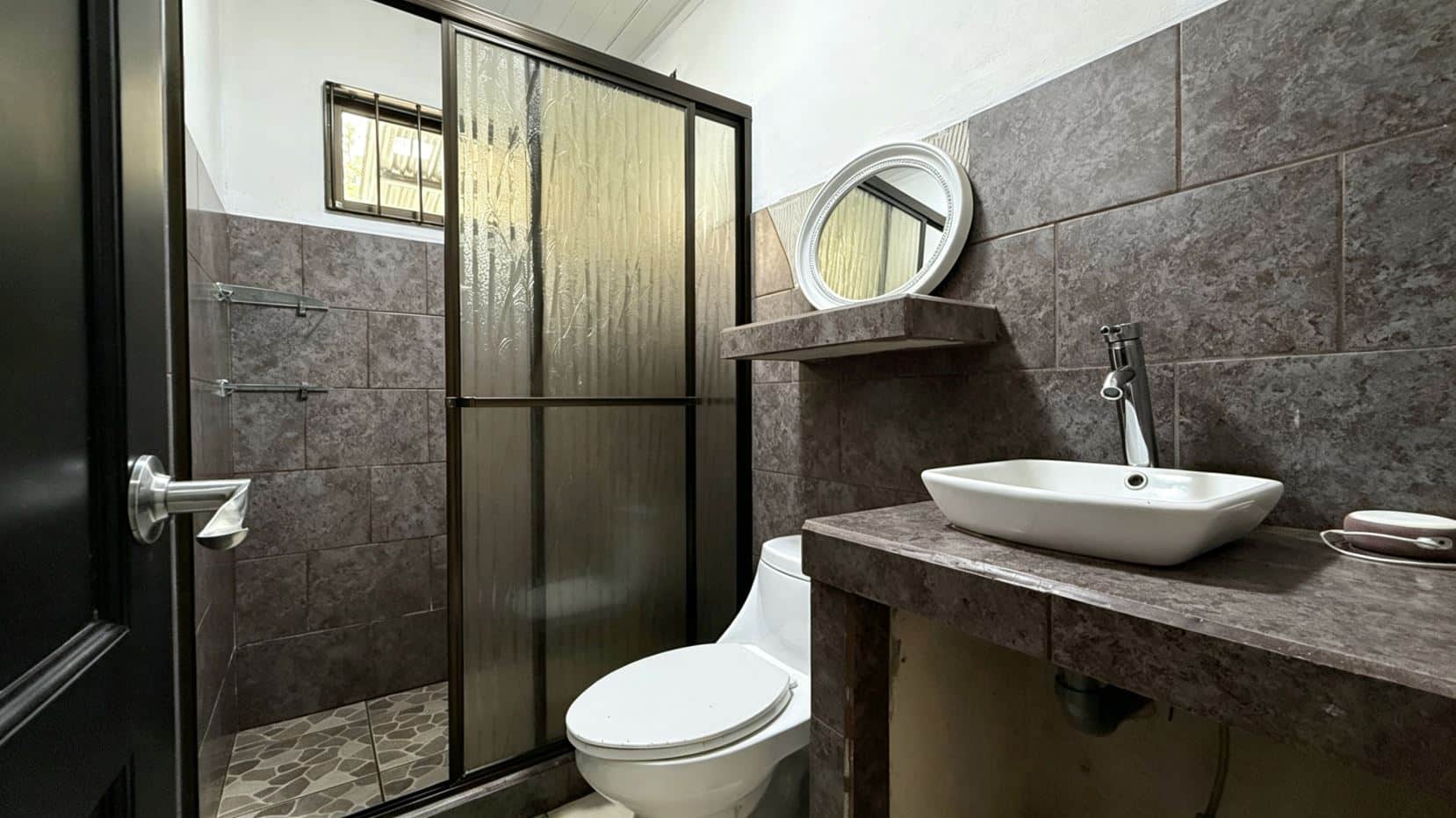 Bathroom with brown tile walls, frosted glass shower enclosure, white vessel sink on a brown countertop, round mirror, and a white toilet nearby.