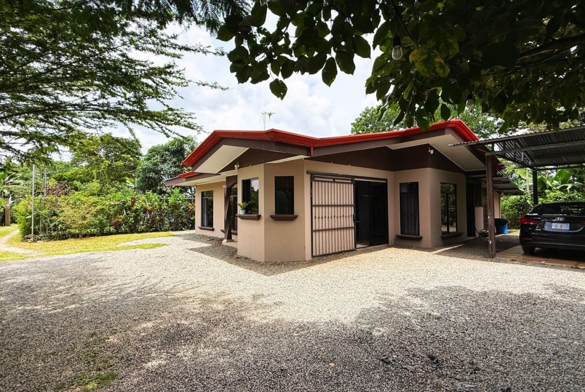 Single-story beige house with a red roof, gravel driveway, and a carport with a black sedan parked beneath it.