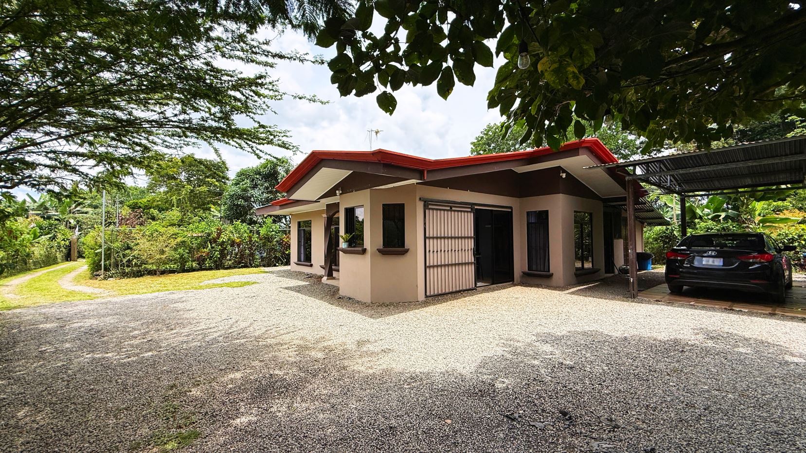 Single-story beige house with a red roof, gravel driveway, and a carport with a black sedan parked beneath it.
