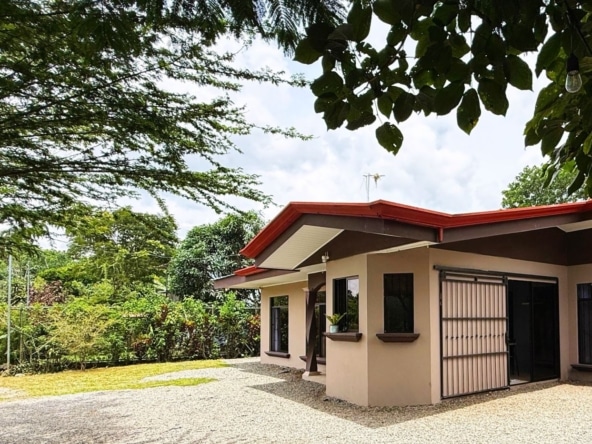 Single-story beige house with a red sloped roof, metal security doors, and a gravel driveway surrounded by lush greenery and trees