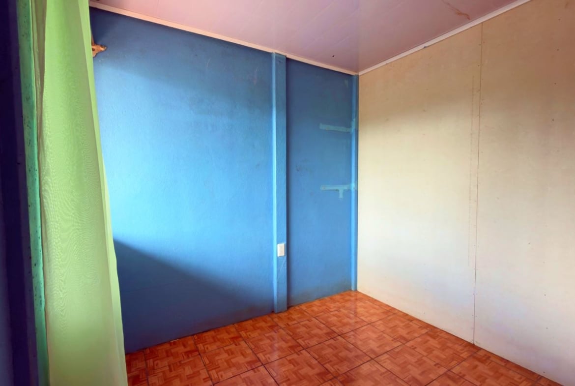 Empty corner of a room with blue walls, a green curtain on the left, brown tiled floor, and a beige wall on the right.