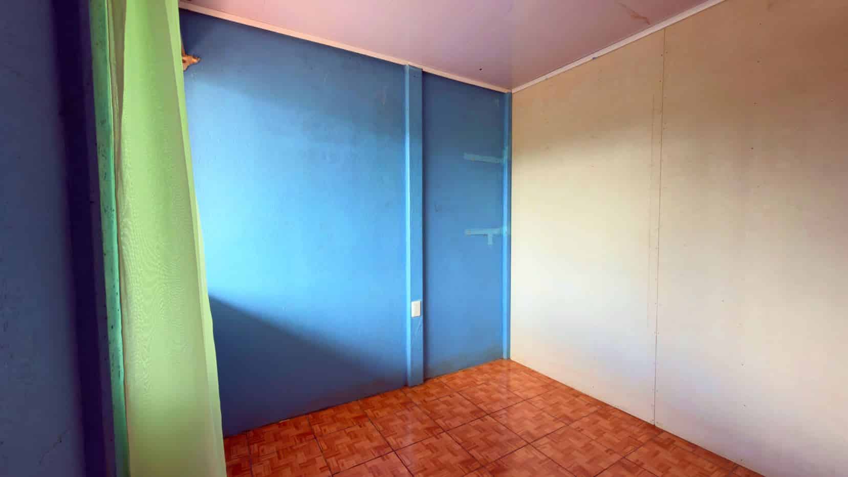 Empty corner of a room with blue walls, a green curtain on the left, brown tiled floor, and a beige wall on the right.