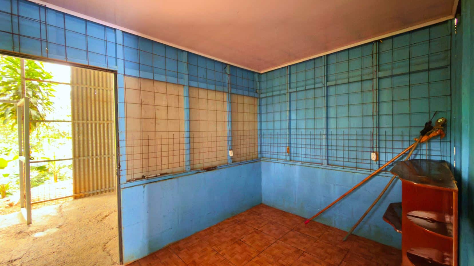 Blue tiled room with metal grid wall panels, a mop leaning on a wooden shelf, and an open door to a sunlit outdoor area.