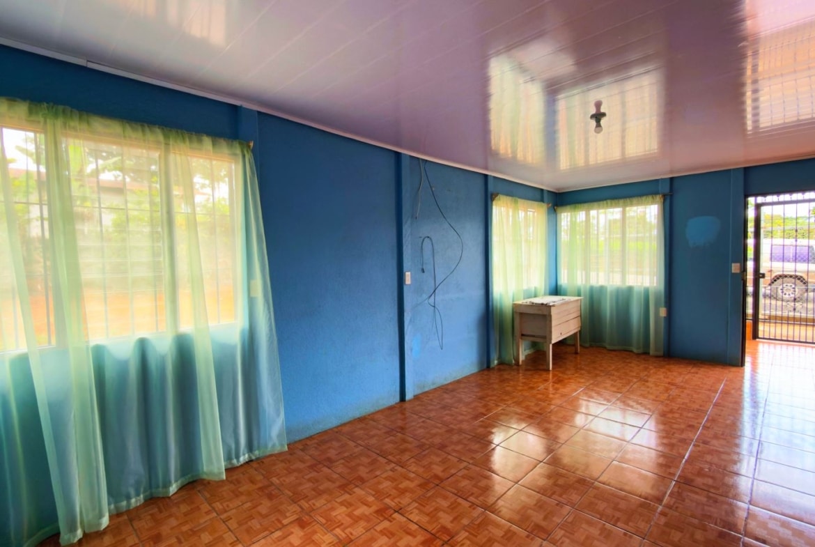 Bright blue living room with turquoise sheer curtains, brown-tiled floor, and a glossy lavender ceiling; sunlight filters through the windows and door opens outside.