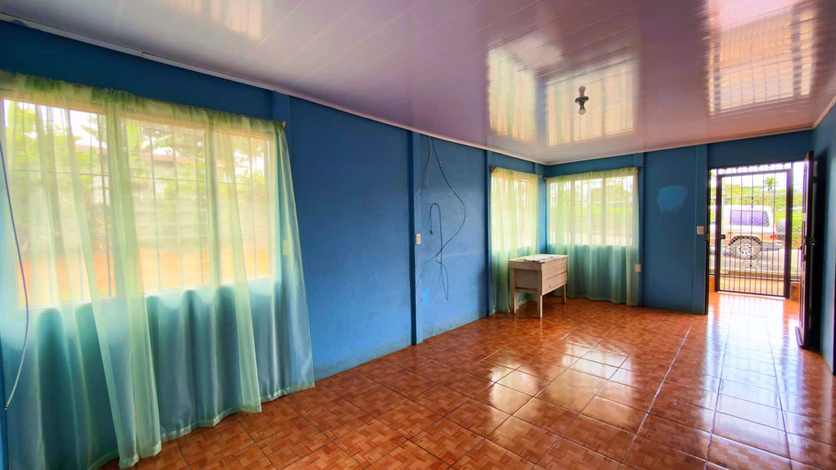 Bright blue living room with turquoise sheer curtains, brown-tiled floor, and a glossy lavender ceiling; sunlight filters through the windows and door opens outside.