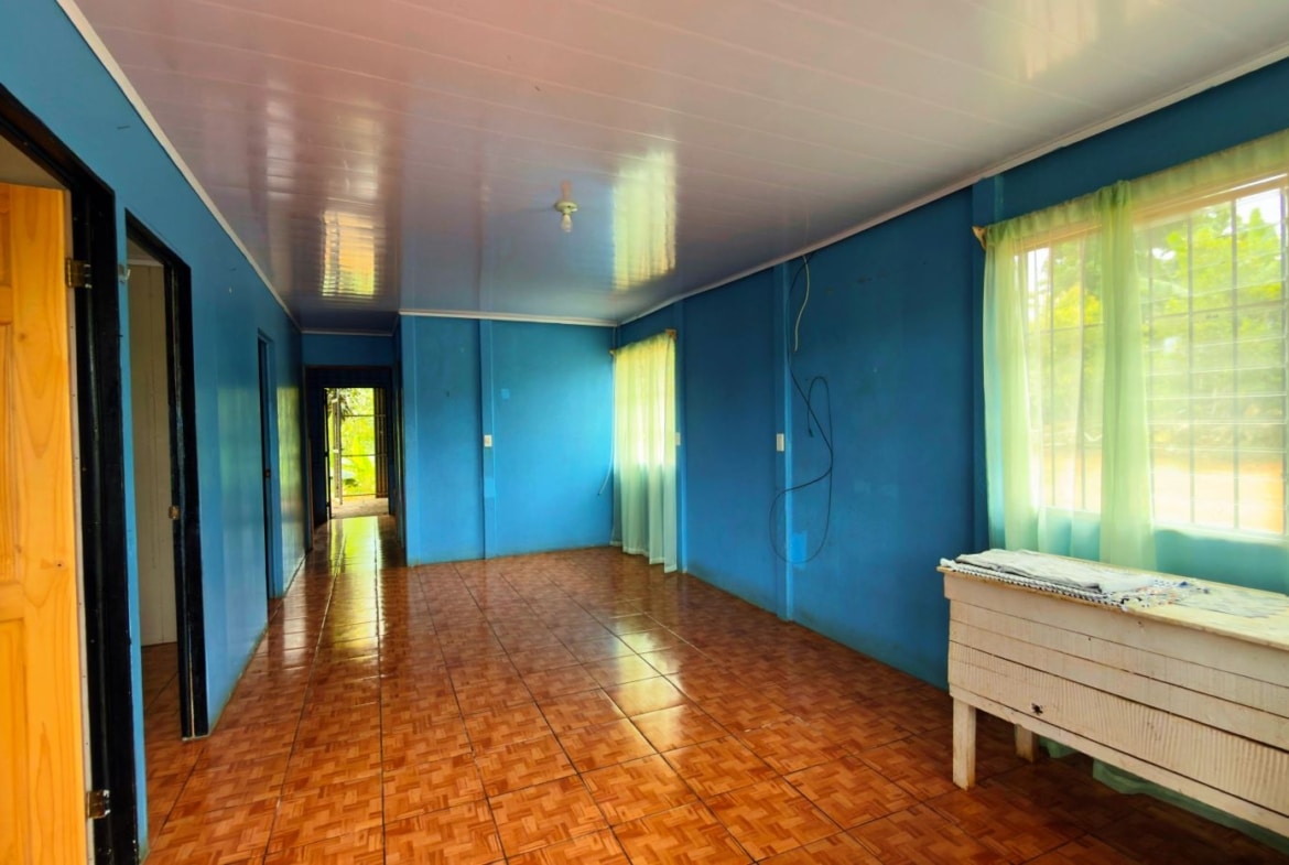 Blue-painted hallway with brown tiled floor, open doors on the left, and large windows with pale green curtains on the right; a white wooden table against the wall.