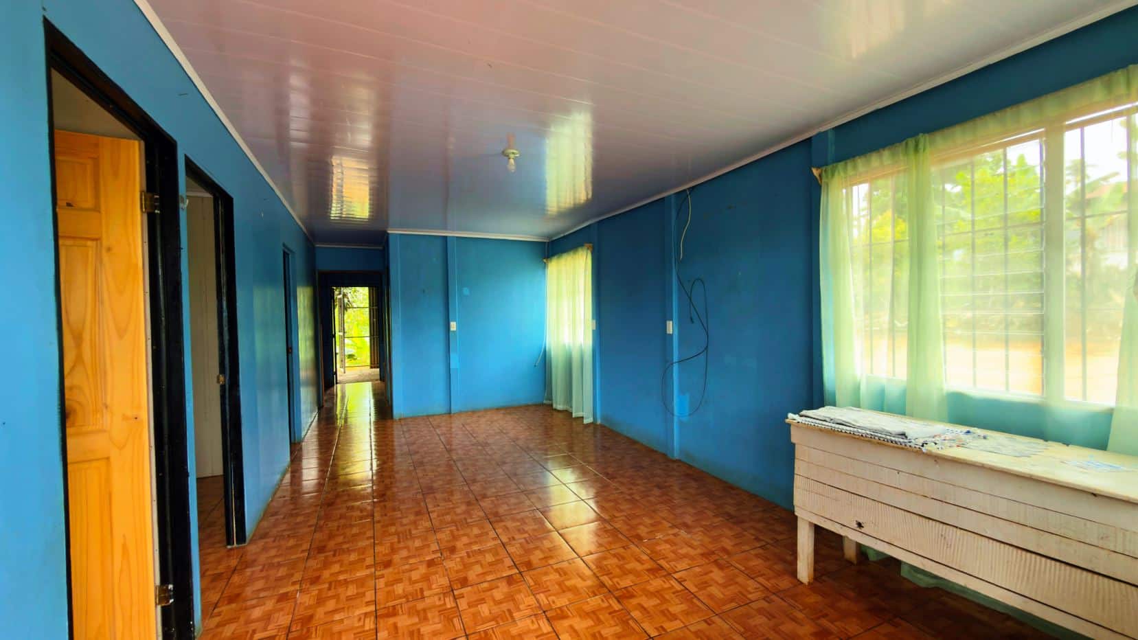 Blue-painted hallway with brown tiled floor, open doors on the left, and large windows with pale green curtains on the right; a white wooden table against the wall.