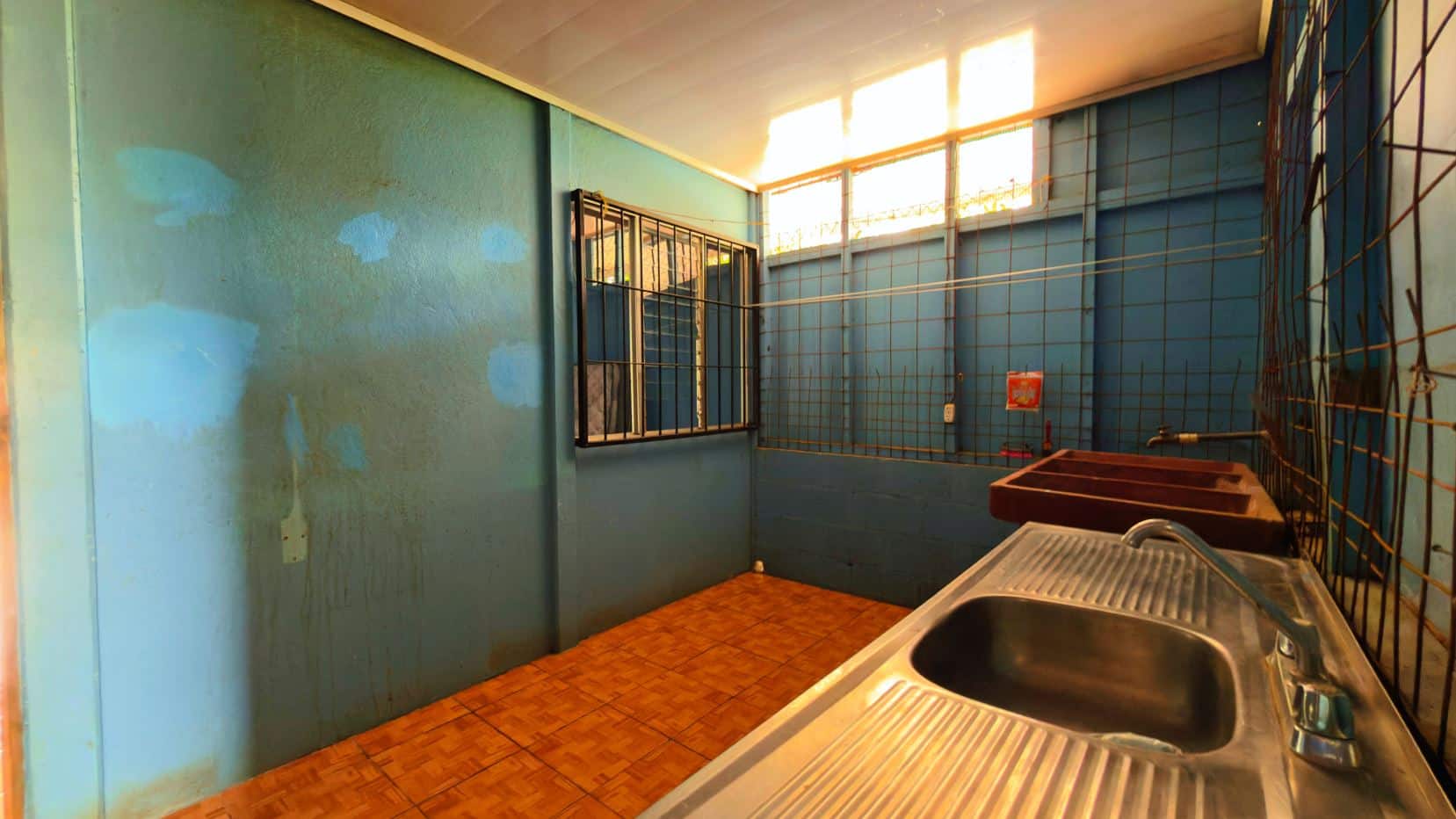 Blue utility room with stainless steel sink in foreground, blue walls, and orange tiled floor. Windows with bars along the back wall.
