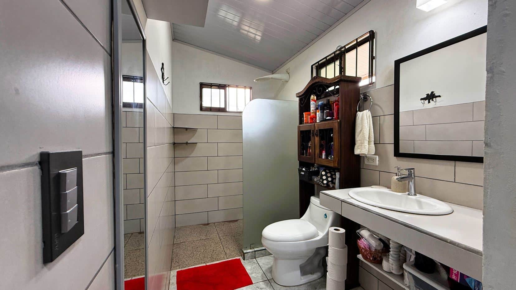 Small bathroom with toilet, sink, mirror, and wooden cabinet against tiled walls and frosted glass divider in the shower area