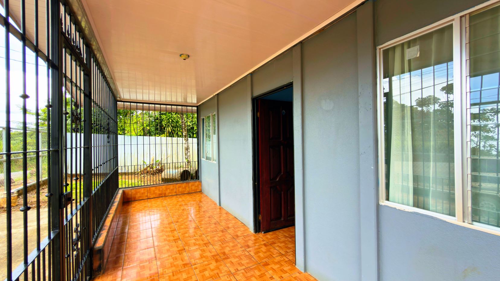 Covered patio area of a blue house with orange tile floor, black metal fence on the left, and an open wooden door on the right.