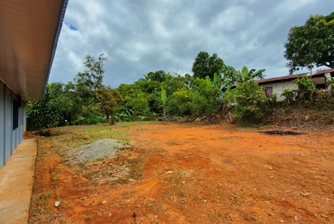 Wide view of an orange dirt lot beside a light blue building with barred windows; dense trees edge the yard.
