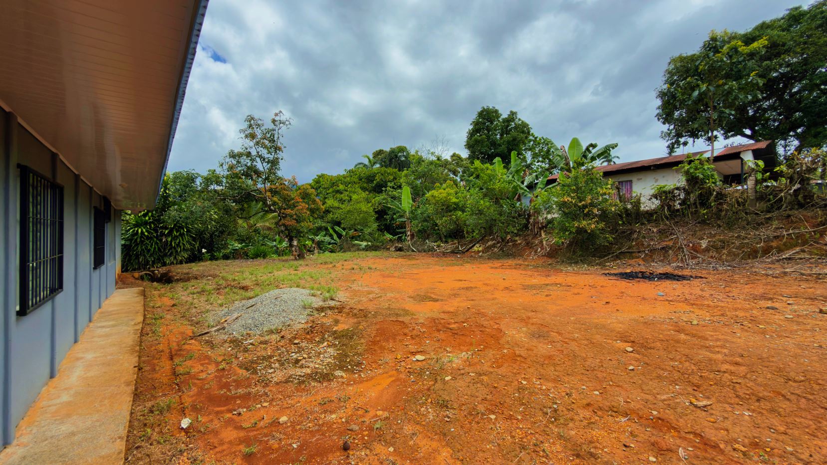 Wide view of an orange dirt lot beside a light blue building with barred windows; dense trees edge the yard.