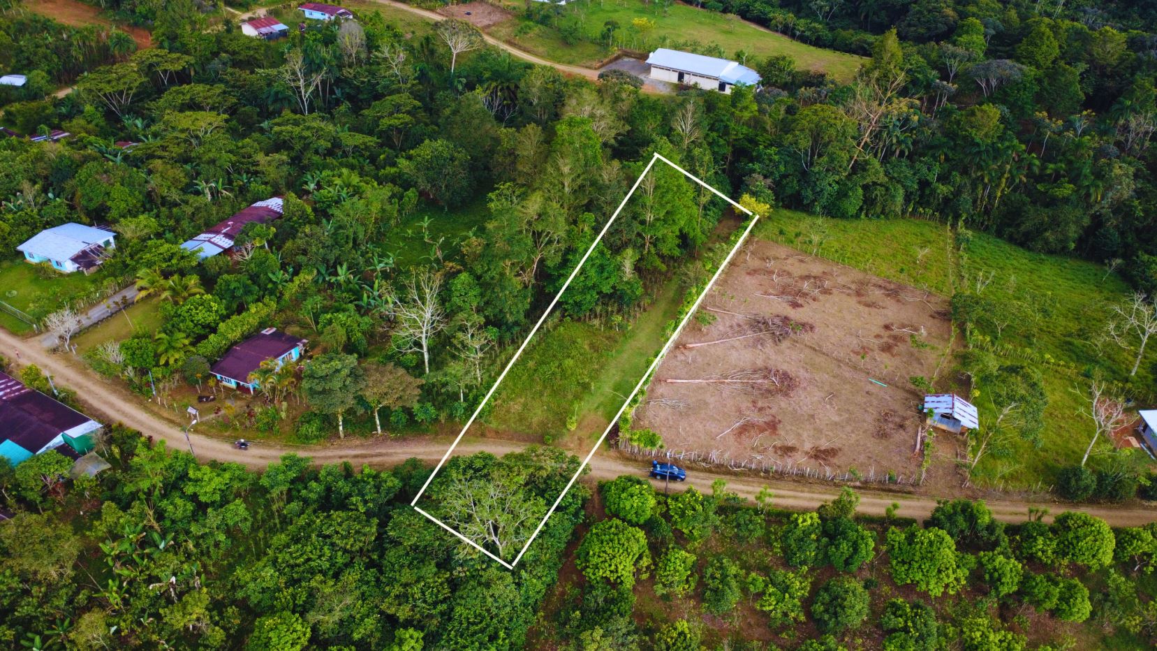 Aerial view of a rectangular plot outlined in white, surrounded by trees and a dirt road with nearby houses.