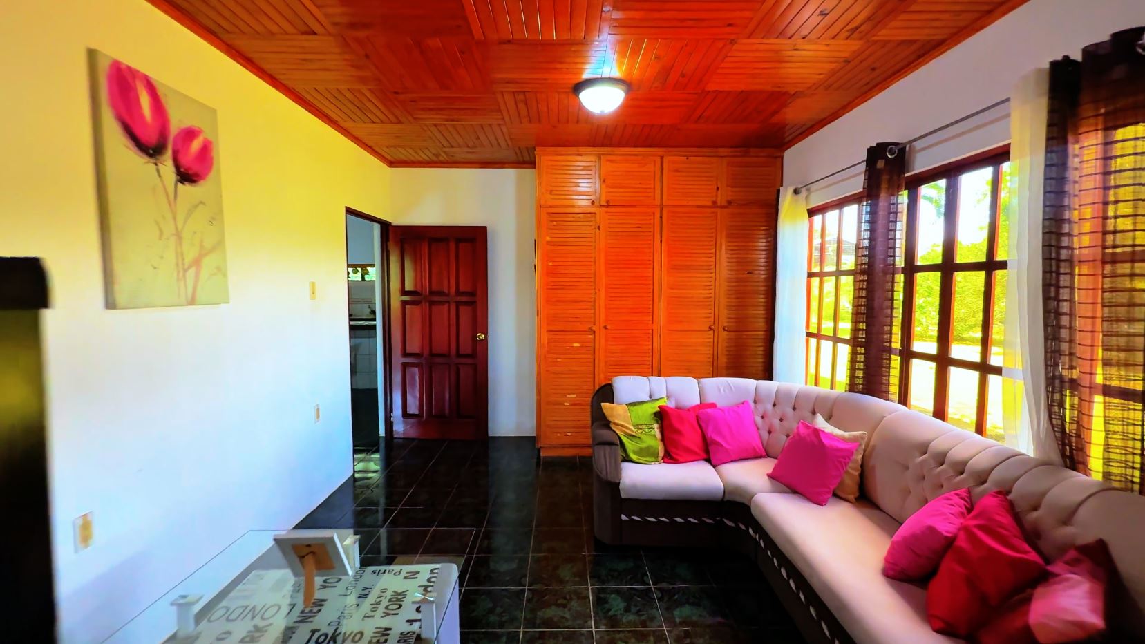 Cozy living room with a beige L-shaped sofa and bright pink and lime cushions, large windows, and a wood-paneled ceiling.