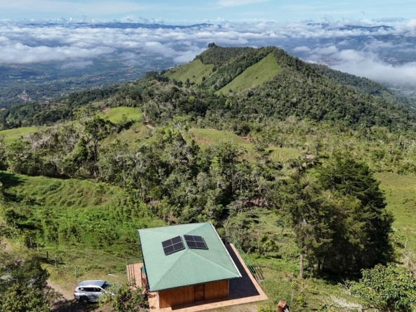 Hilltop cabin with a green roof and solar panels, surrounded by forest and rolling green hills under a cloudy sky.