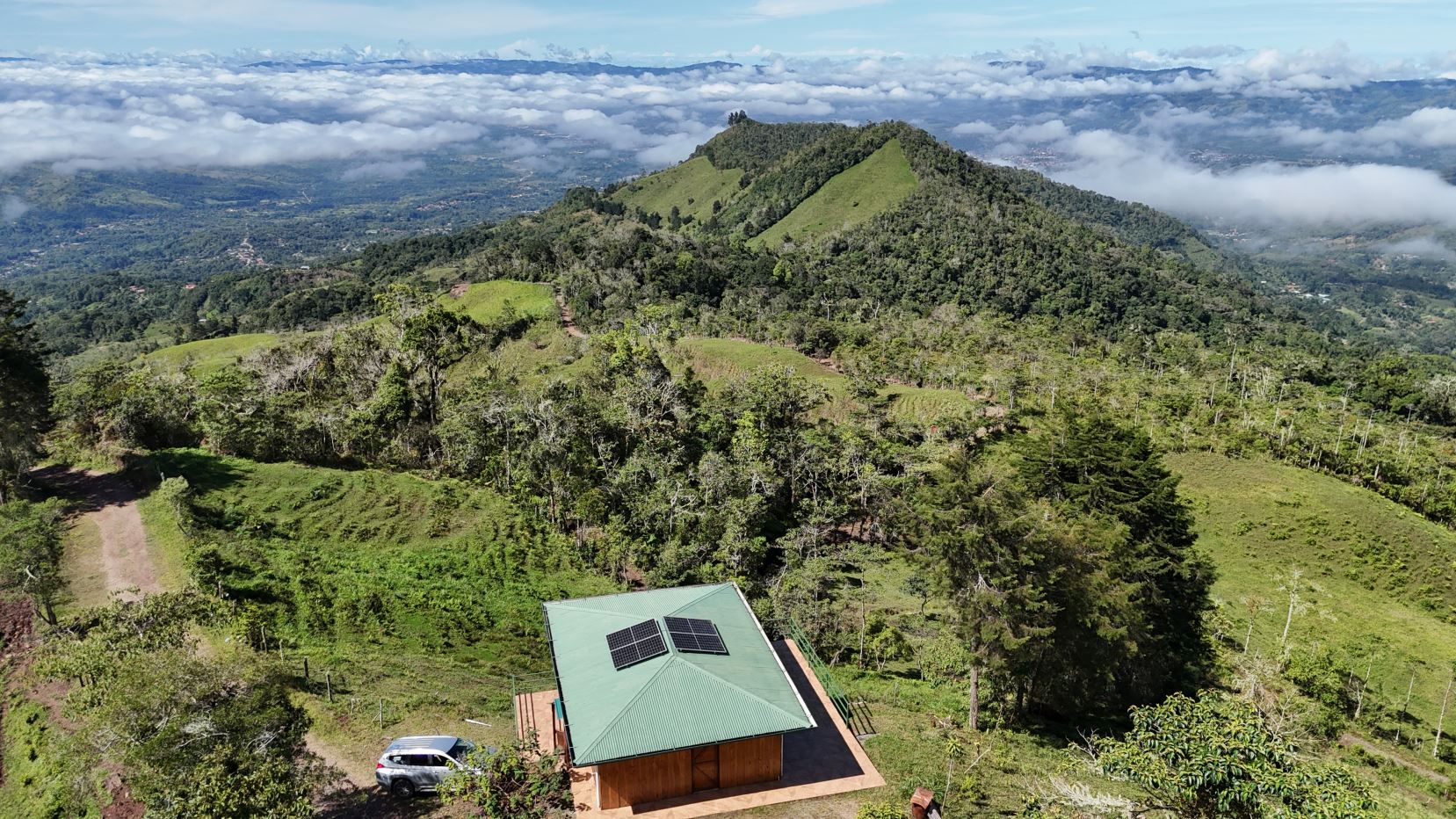 Hilltop cabin with a green roof and solar panels, surrounded by forest and rolling green hills under a cloudy sky.