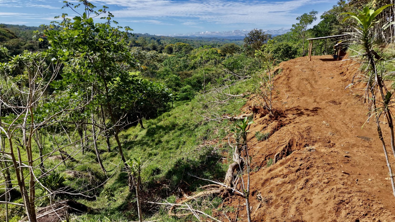 Dirt path along a hill’s edge with exposed soil and a small wooden shelter on the right, overlooking dense green jungle under a blue sky.