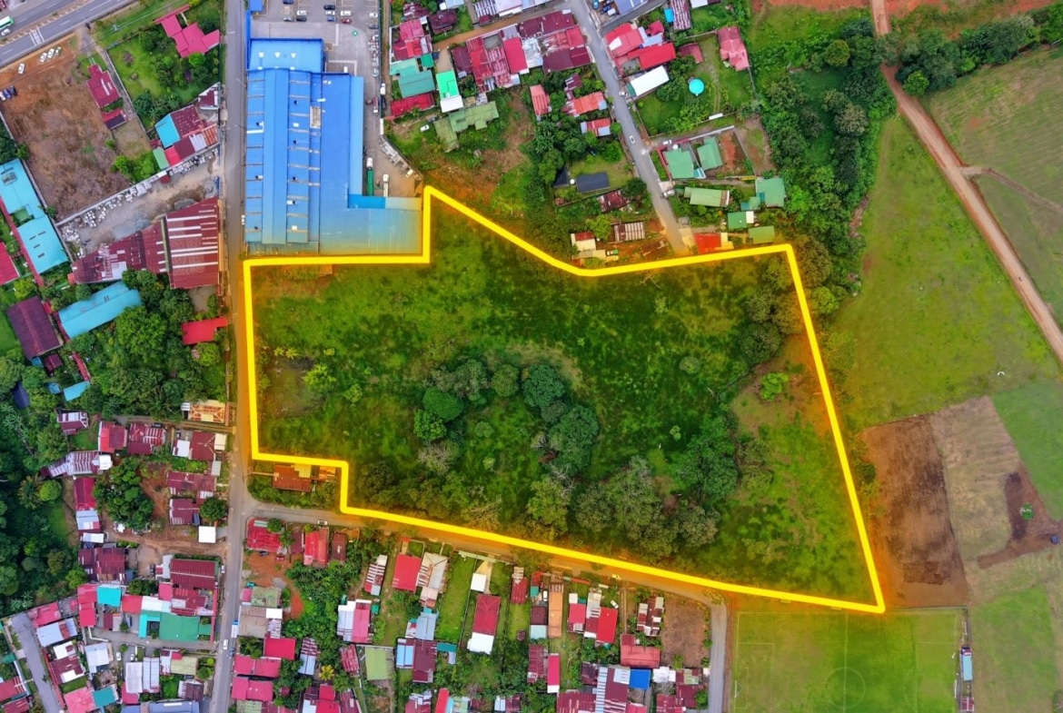 Aerial view of a large grassy lot outlined in bright yellow, bordered by a residential area and open fields behind and to the sides for context.